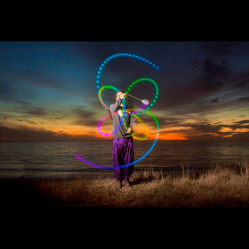 a performer spinning flowtoys LED podpoi with rainbow trails during sunset near a lake